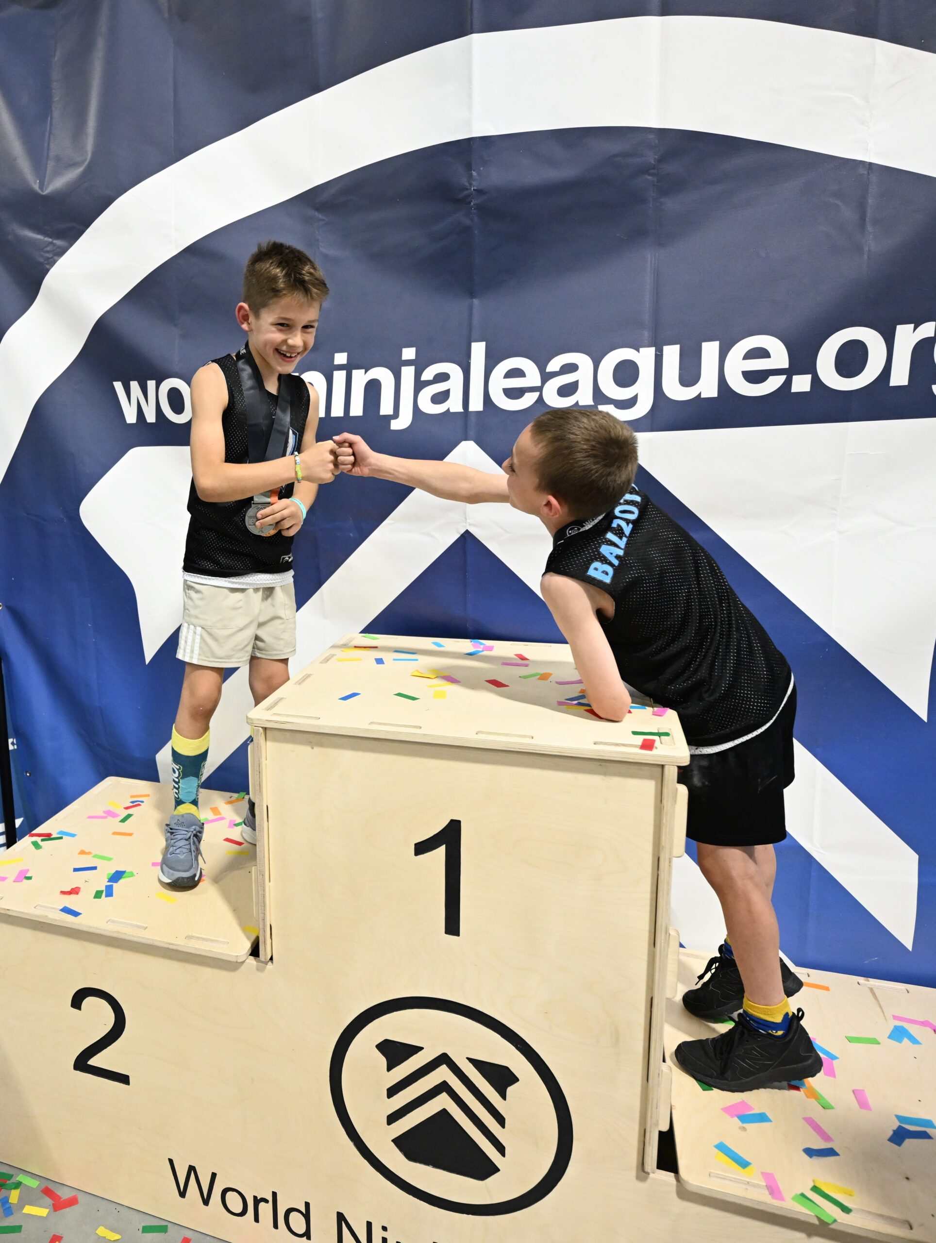 Two smiling young male ninja athletes standing on a podium after an awards ceremony for the World Ninja League Regional Championships congratulate each other