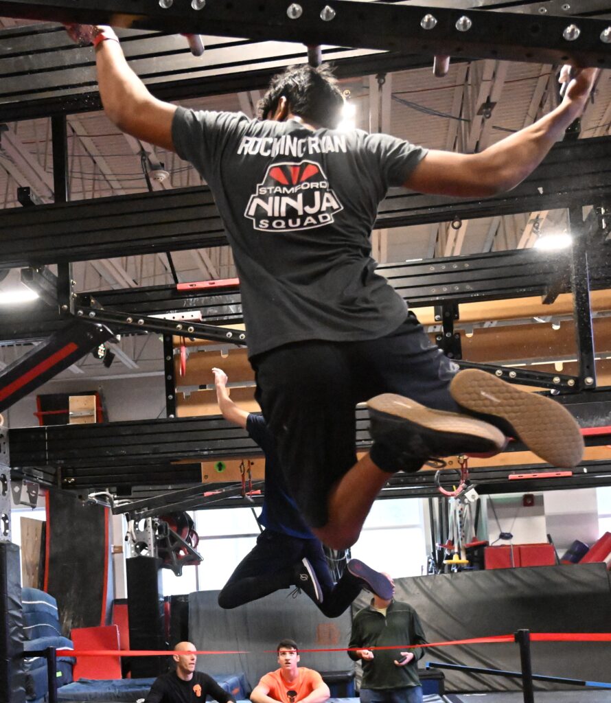 Two male ninja athletes compete in a head to head race at the World Ninja League World Championships. Both athletes are traversing across pegs suspended above the ground.